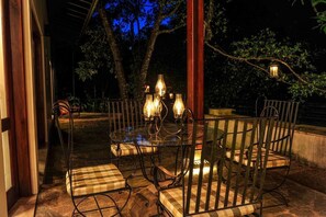 An outdoor dining area with a glass table and metal chairs, surrounded by trees and illuminated by hanging lanterns. - The Weir House (Ulapane)
