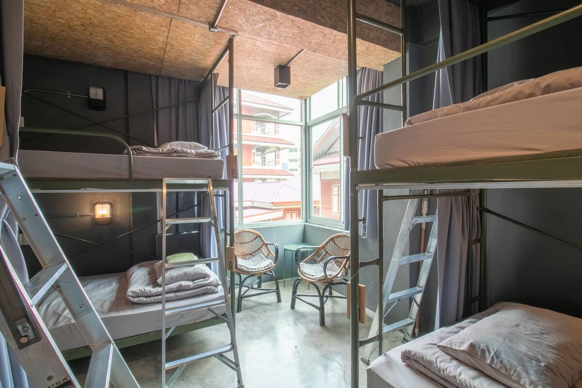 A dormitory room with bunk beds, a ladder, two wicker chairs, and a window with a view of buildings.