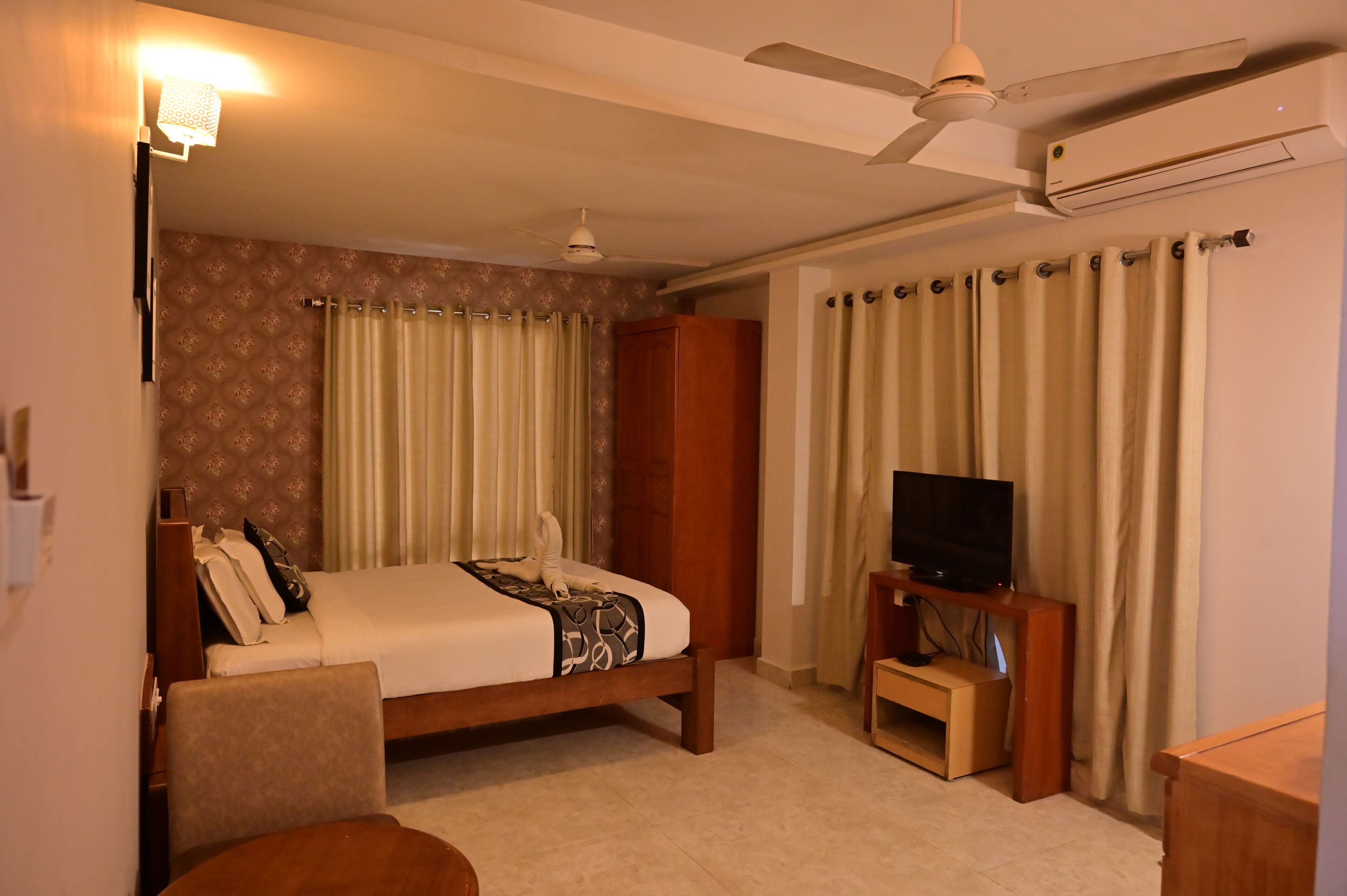 A hotel room with a bed, a TV on a wooden stand, a ceiling fan, and an air conditioning unit.