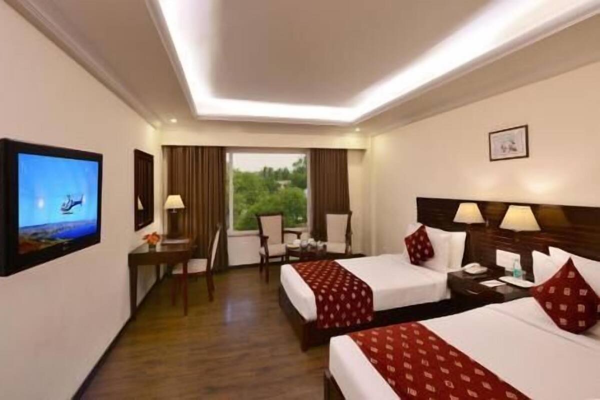 A hotel room with two beds, a TV, a desk, and a window with a view of greenery.
