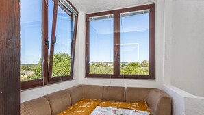 A corner room with a sofa and a table, large windows with wooden frames, and a view of greenery and buildings.