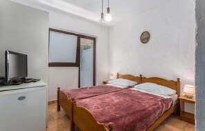 A bedroom with a bed, a TV, a window with blinds, a bedside table with a lamp, and a wall clock.