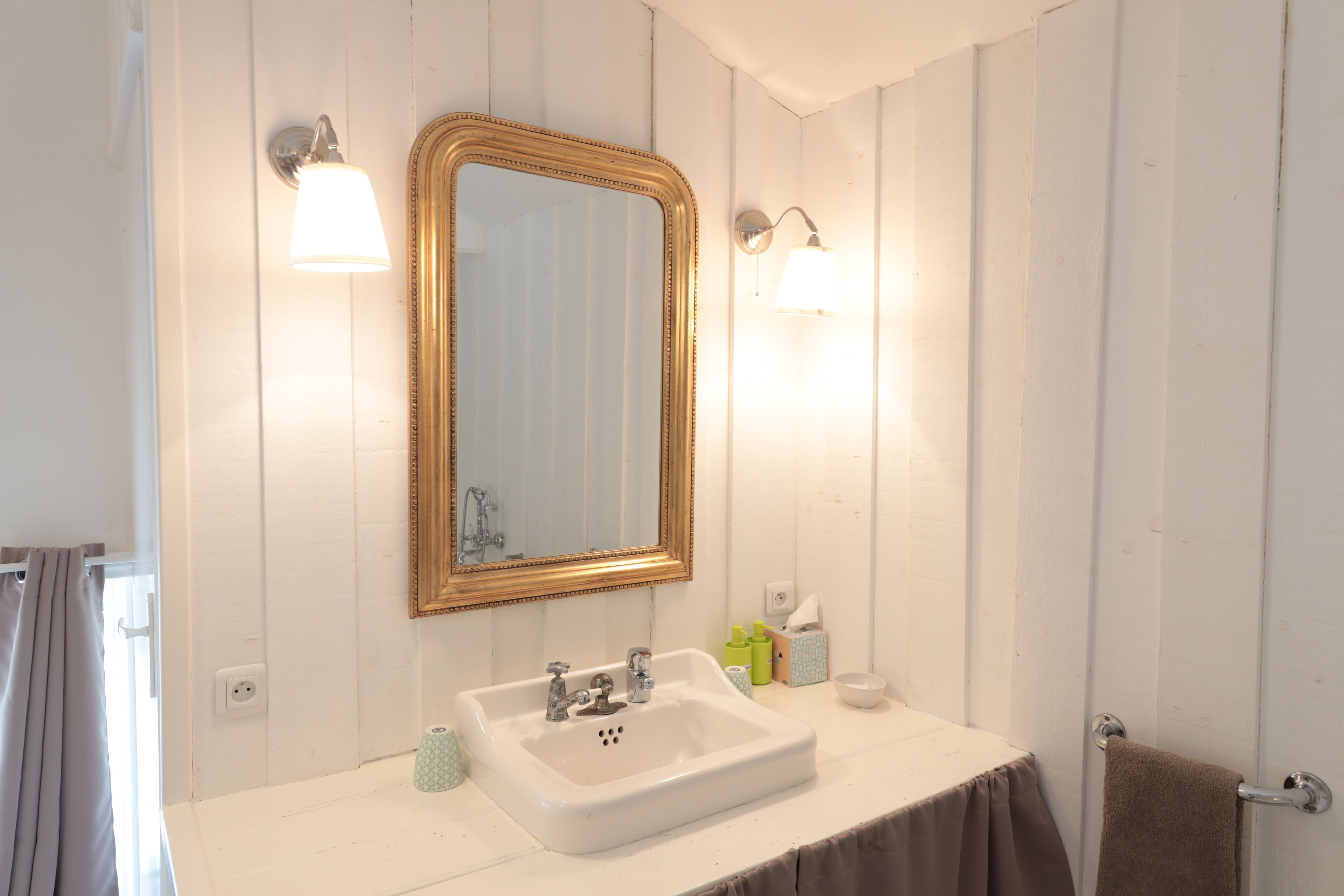 Une salle de bain avec un lavabo blanc, un miroir doté d’un cadre doré et deux appliques murales.