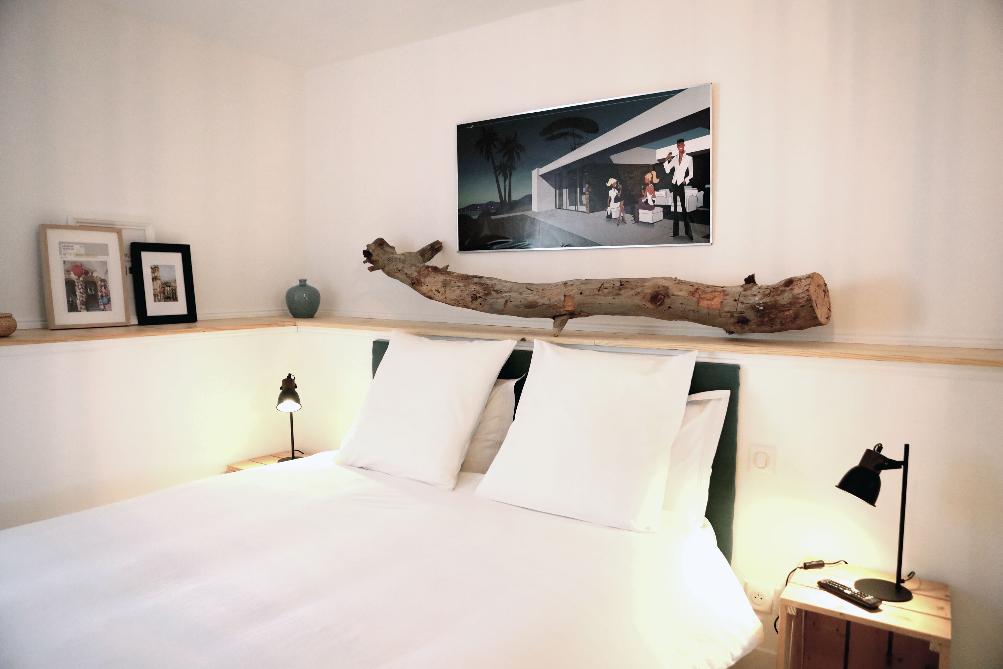 Una cama bien hecha con sábanas blancas, cabecera y dos lámparas de noche. Encima de la cama, en la pared, cuelga una fotografía enmarcada.
