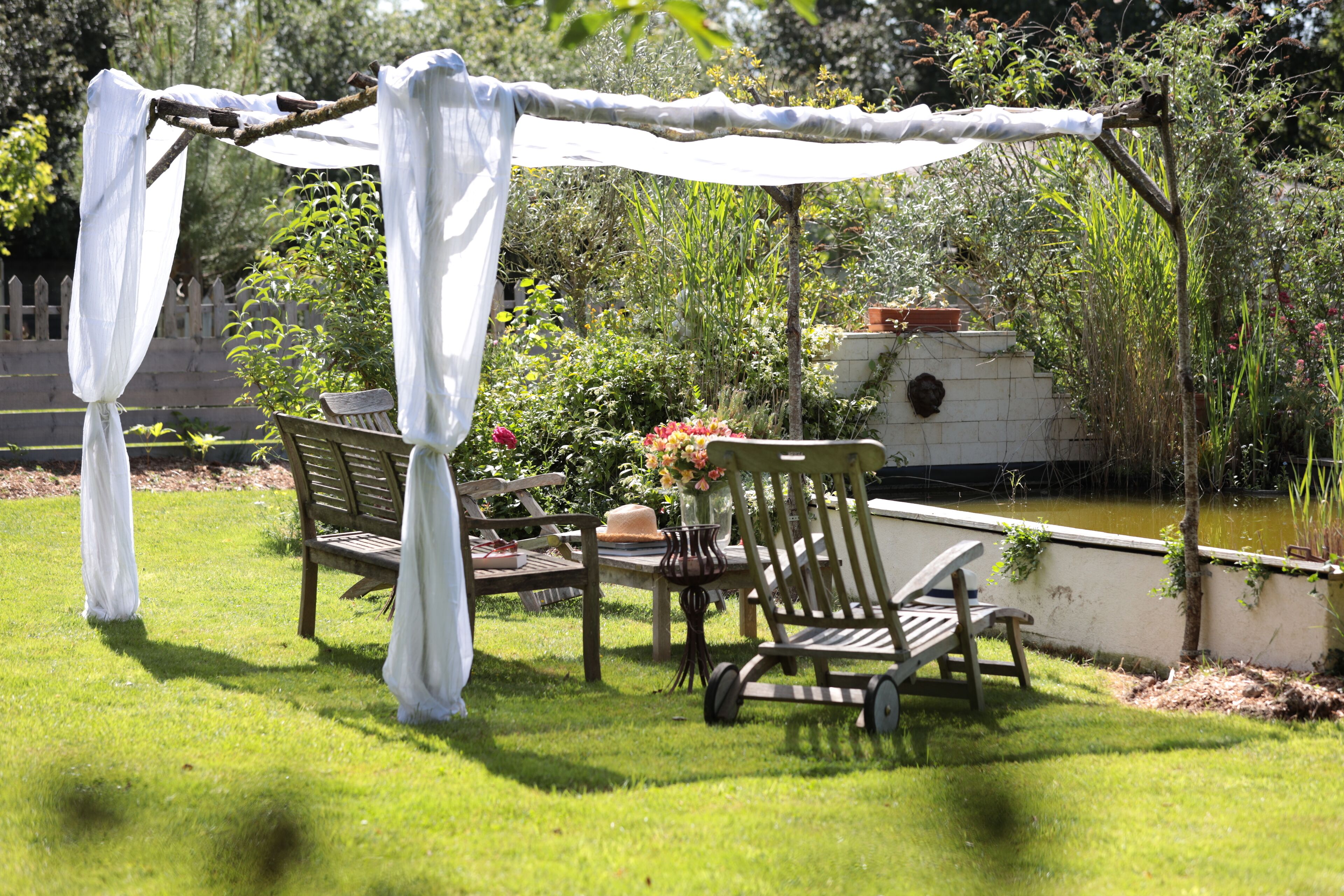 An outdoor seating area with a table and chairs under a canopy, surrounded by greenery and a water feature.