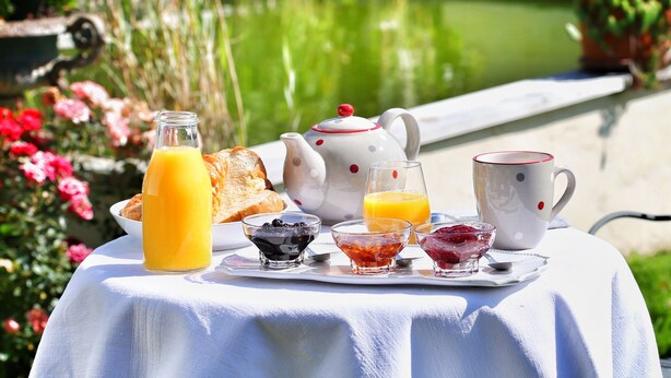 Un petit-déjeuner composé de toasts, de jus de fruits et de thé, servi sur une table près d’un étang.