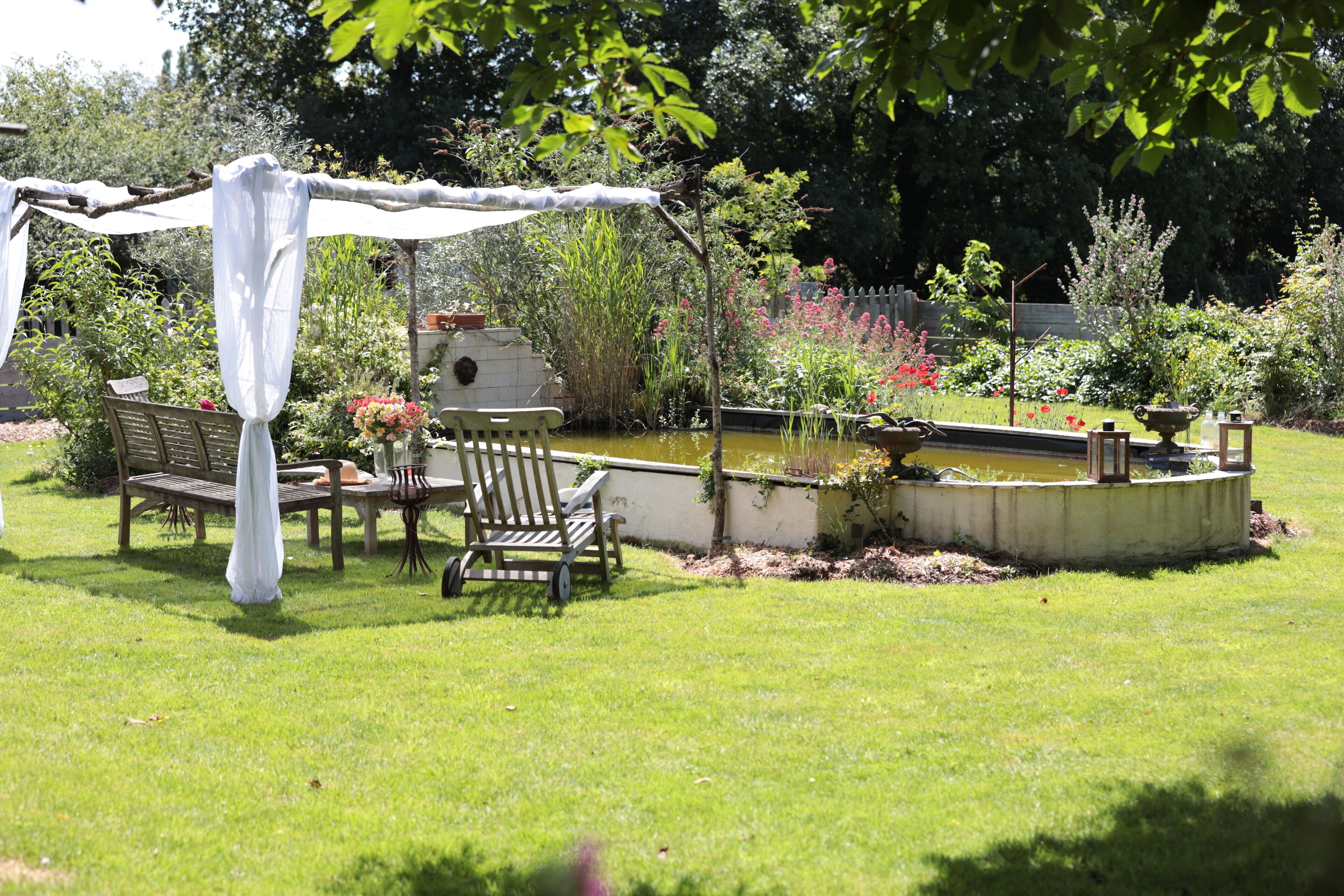 Een tuin met een houten bank, een tafel en een pergola met witte stof.
