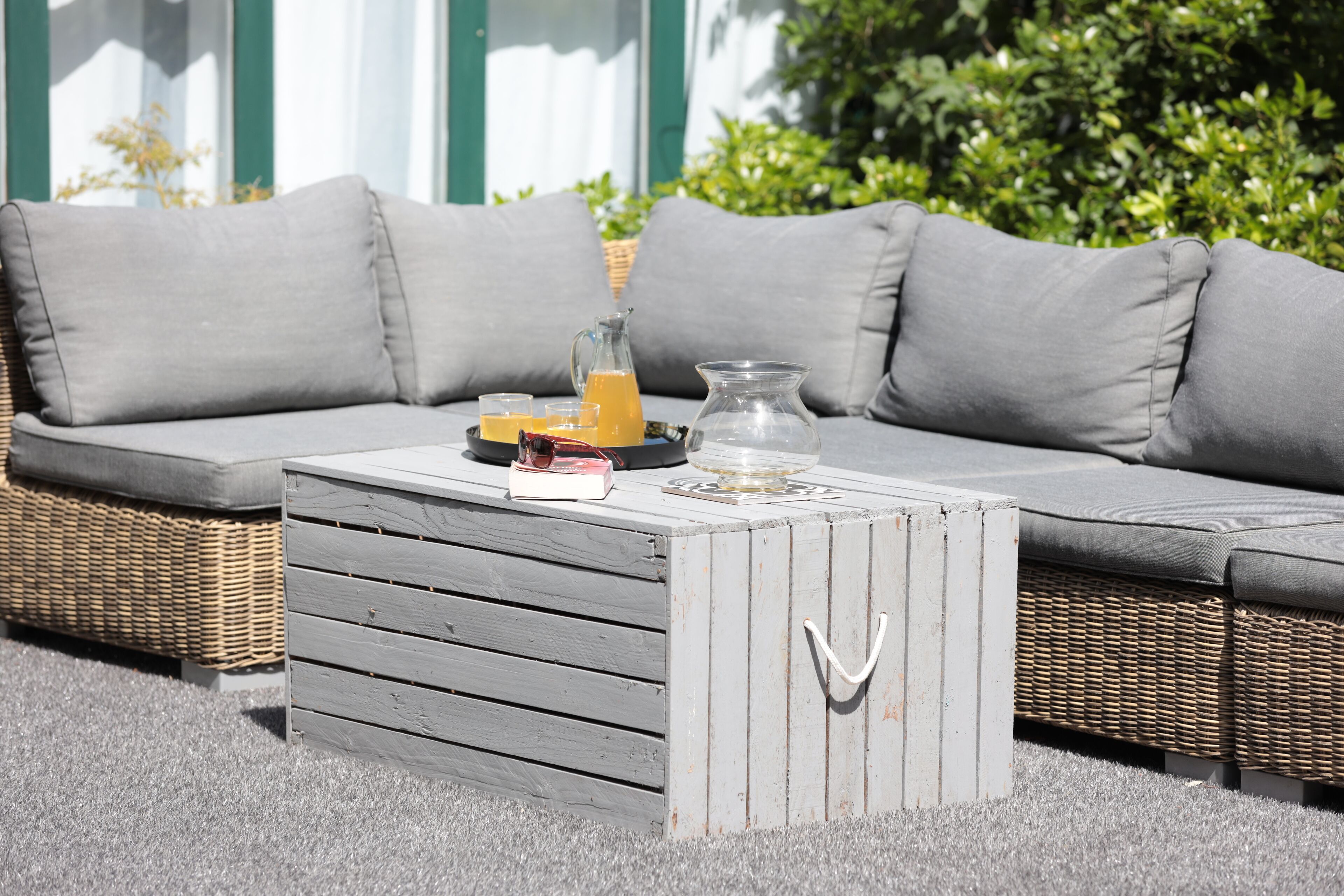 Un sofá gris con cojines, una mesa de centro de madera con una jarrón de vidrio y jugo de naranja, y una cesta de mimbre.