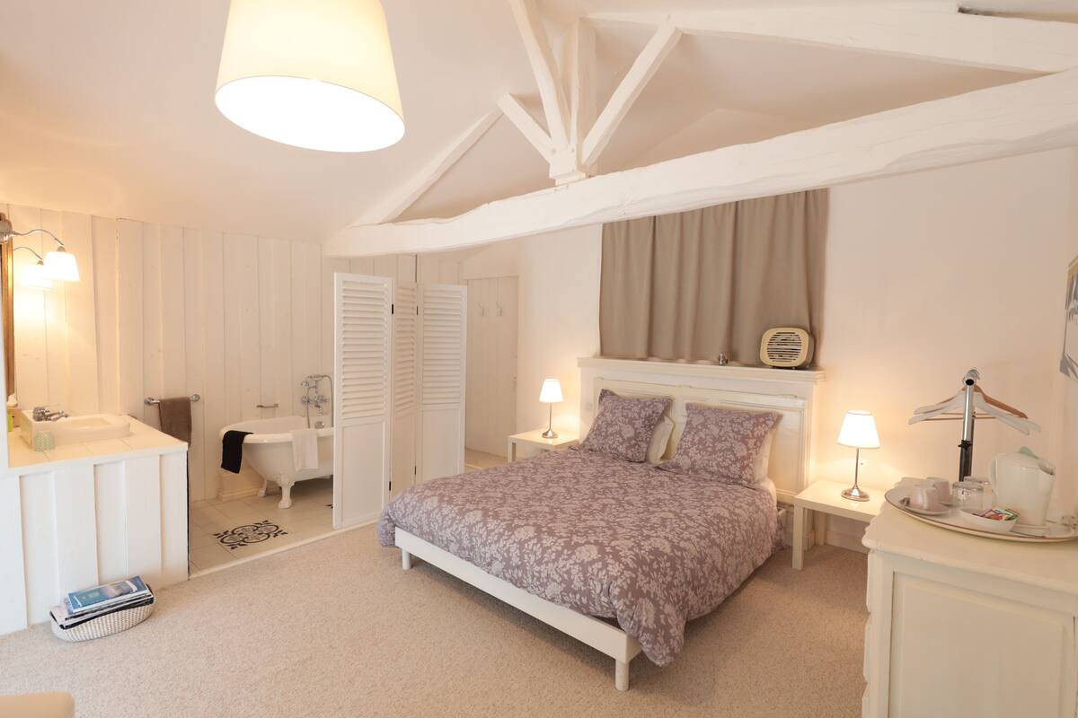 Une chambre à coucher comprenant un lit, des tables de chevet, une commode, un meuble à tiroirs, un miroir, une fenêtre avec des rideaux et un plafond avec des poutres apparentes.