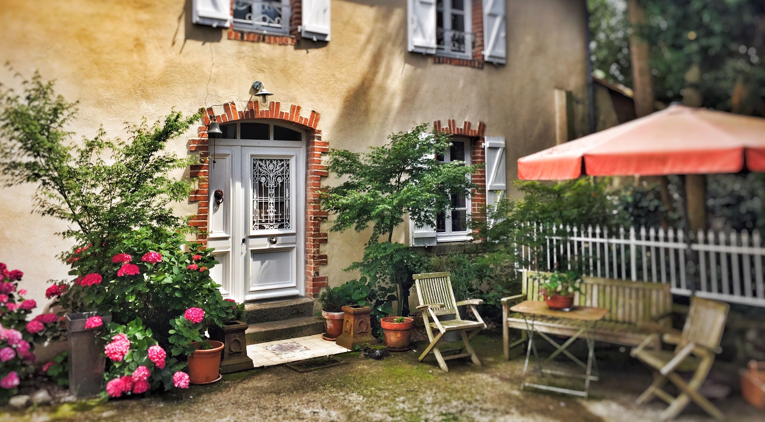 Une charmante maison avec une porte blanche, un petit porche et un parasol rouge. On y trouve des plantes en pot et une chaise en bois à l’extérieur.