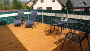 Une terrasse en bois, avec des chaises et une table en métal, entourée d’un mur recouvert de carreaux verts et blancs.