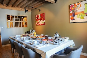Une table de petit-déjeuner dressée avec différents plats et boissons, une fenêtre avec des stores et un tableau accroché au mur.