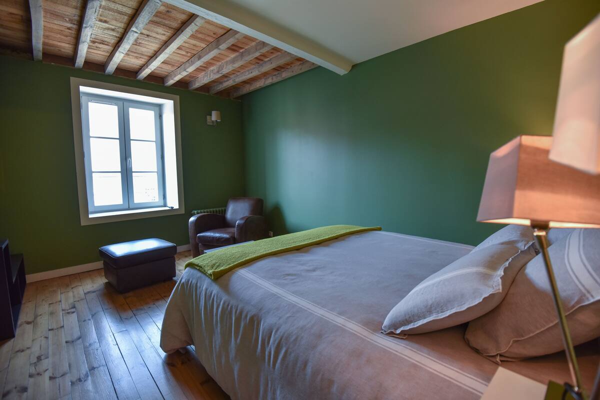 A bedroom with a wooden ceiling, a large bed with pillows, a chair, a window, and a nightstand.