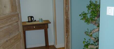 Un couloir étroit au plancher de bois, une petite table en bois avec une bouilloire et une tasse, et une plante en pot.