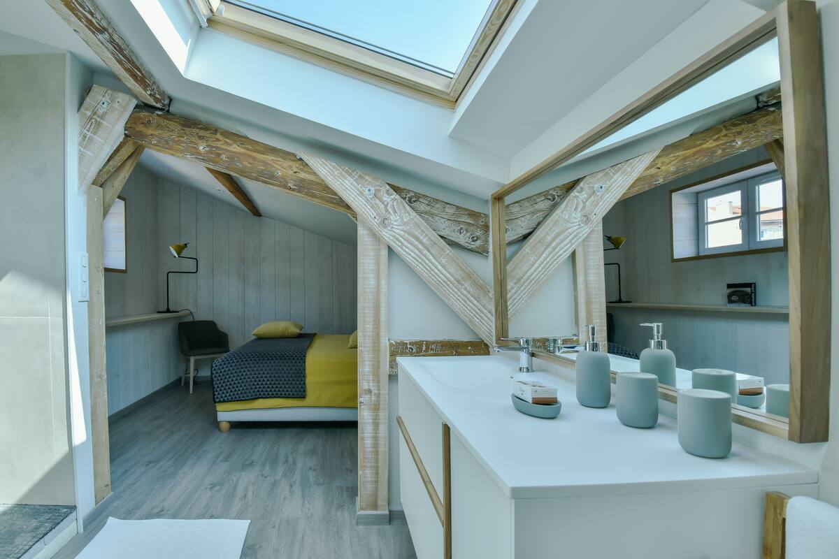 A modern bedroom with a skylight, a bed with a yellow blanket, a white vanity with multiple mirrors, and a wooden ceiling.