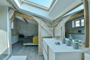 Une chambre moderne avec une lucarne, un lit recouvert d’une couverture jaune, une coiffeuse blanche dotée de plusieurs miroirs et un plafond en bois.