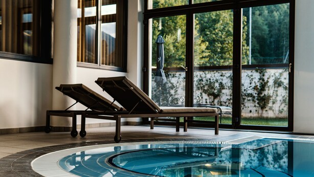 Piscina interior moderna con dos tumbonas y ventanas amplias que ofrecen vistas a la vegetación exterior.