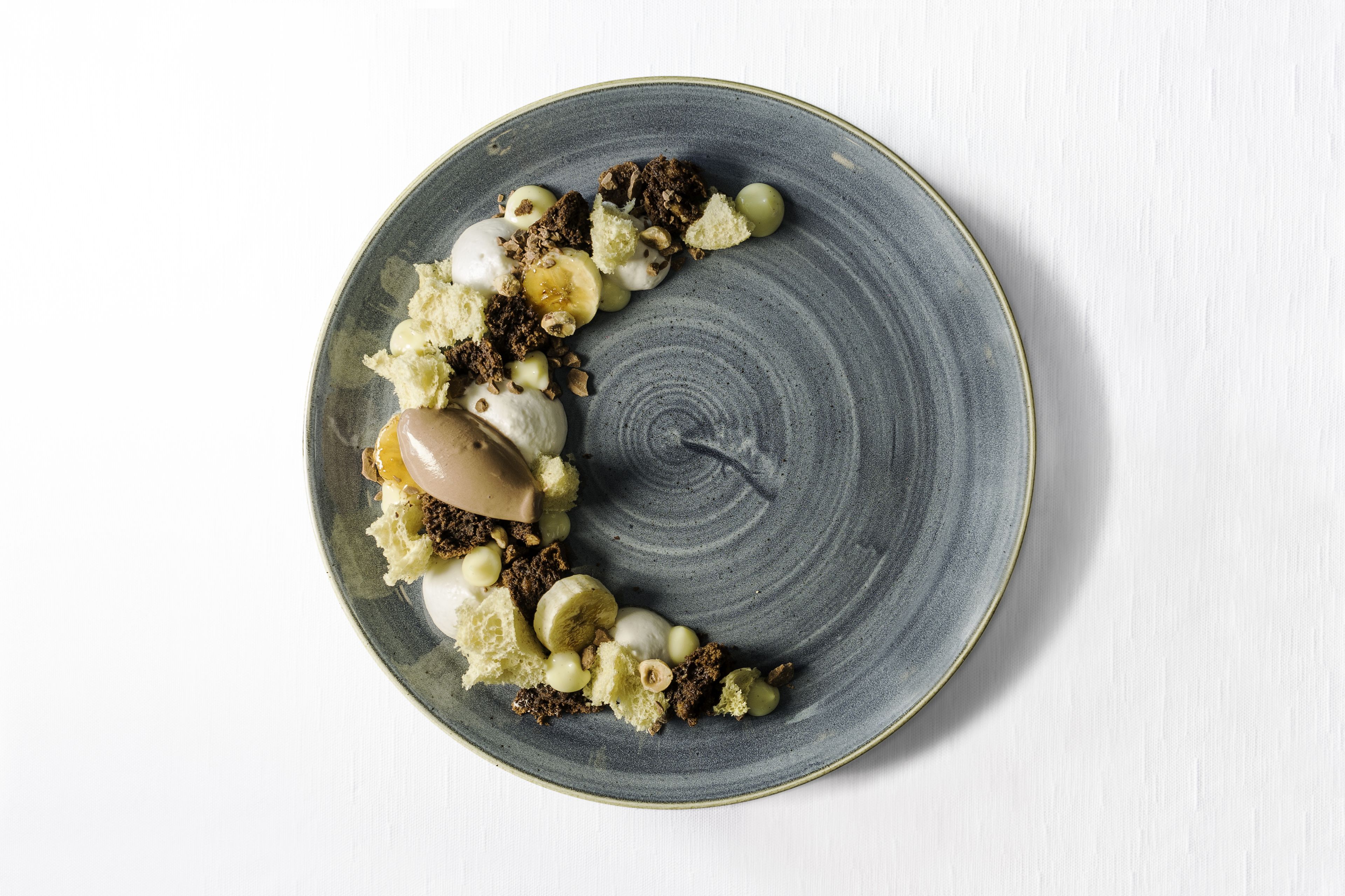 Une assiette ronde en céramique, ornée d’un motif texturé, et contenant divers aliments, notamment une part de gâteau, des noix et des raisins verts.