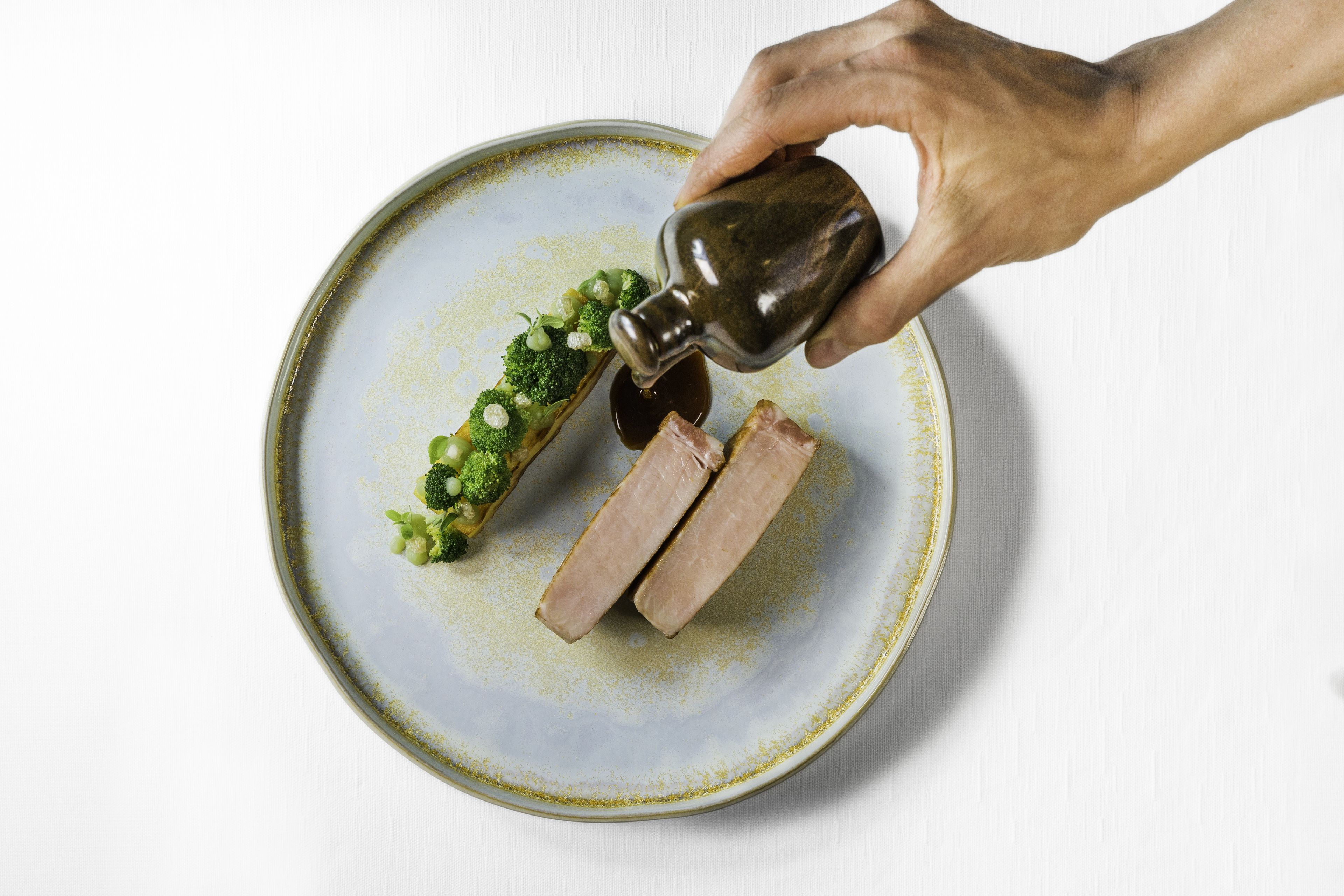Una mano sirviendo salsa sobre un plato con carne y brócoli.