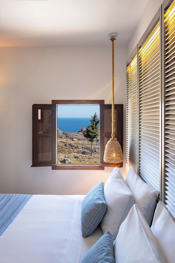A bedroom with a bed, pillows, a hanging light fixture, and a window with a view of the sea.