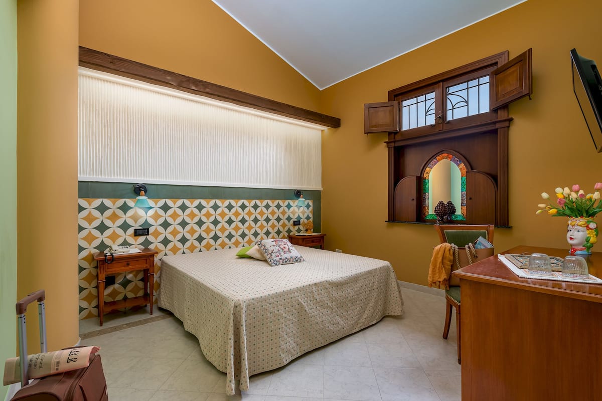 Una habitación con cama, escritorio, silla, ventana con persianas y una ventana arqueada decorativa.