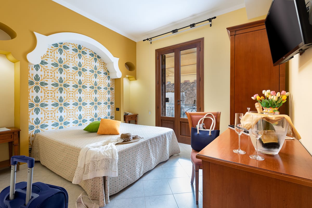 Habitación de hotel con cama, un escritorio con un florero, una maleta y un televisor en la pared.