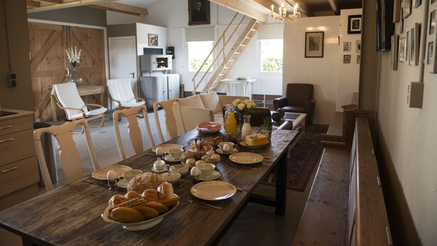 A dining area with a long wooden table set for breakfast, surrounded by white chairs, with a staircase and kitchen in the background. - B&B Logeren bij van Heeren (Made)