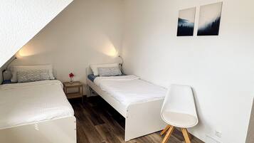 A small, triangular attic bedroom with two beds, a nightstand, a chair, and two wall-mounted pictures.
