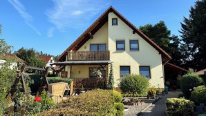 Ein zweistöckiges Haus mit Balkon, umgeben von gepflegten Gärten und blauem Himmel.