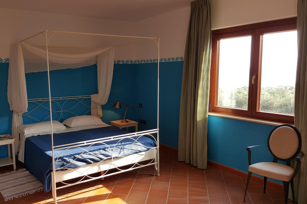 Ein Schlafzimmer mit einem Himmelbett, einem Nachttisch mit Lampe, einem Sessel und einem Fenster mit Vorhängen.