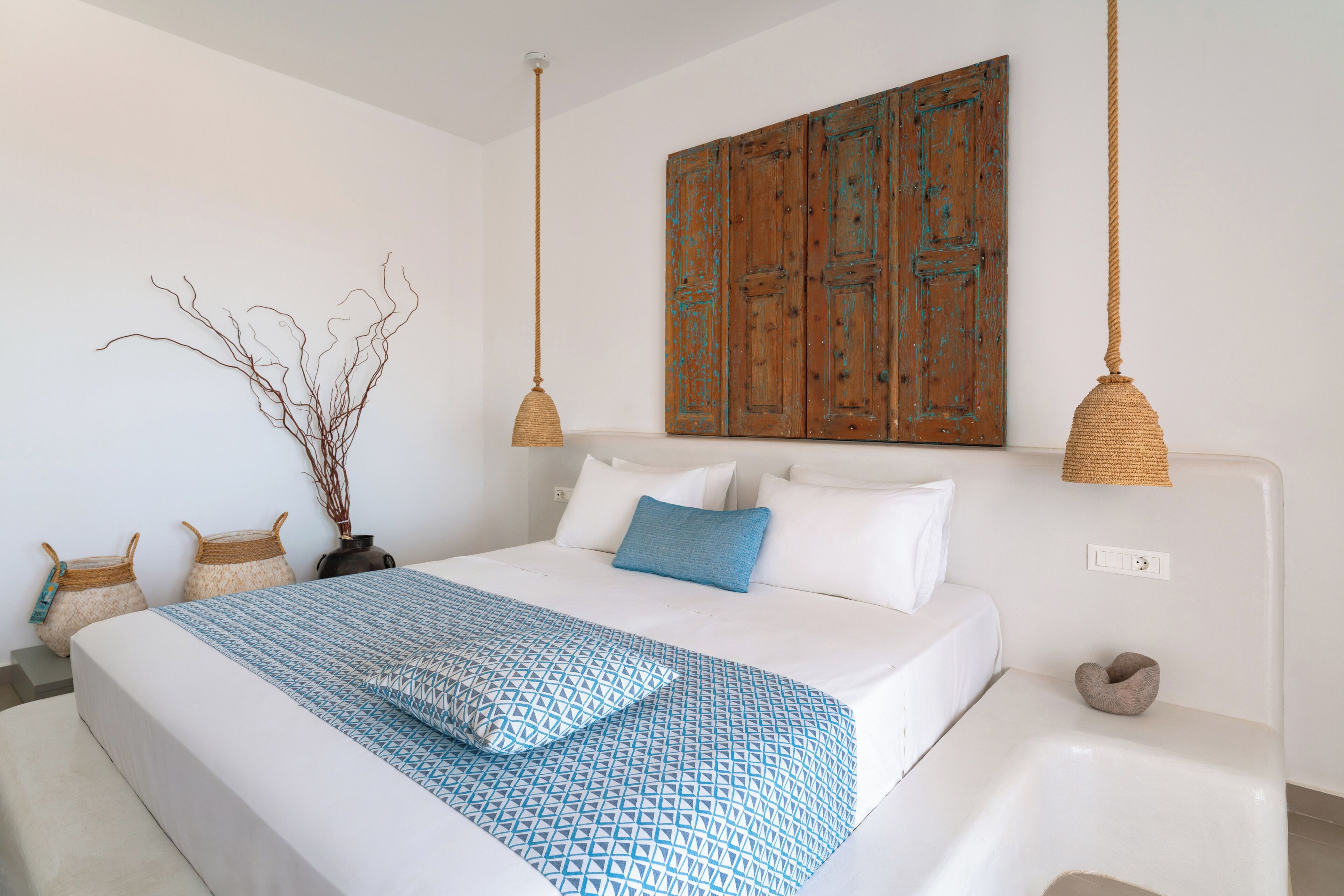 A bedroom with a large bed, white bedding, a blue patterned throw, a wooden door, and two hanging light fixtures.