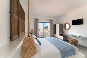 A modern hotel room with a large bed, a desk, a TV, and a view of the outdoors. - Lindos Portes Suites - Adults Only (Rhodes)