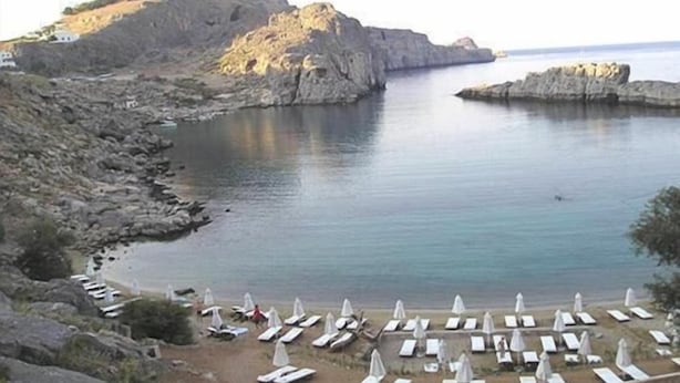 A coastal beach with rocky cliffs, a calm sea, and rows of sun loungers with umbrellas. - Lindos Portes Suites - Adults Only (Rhodes)