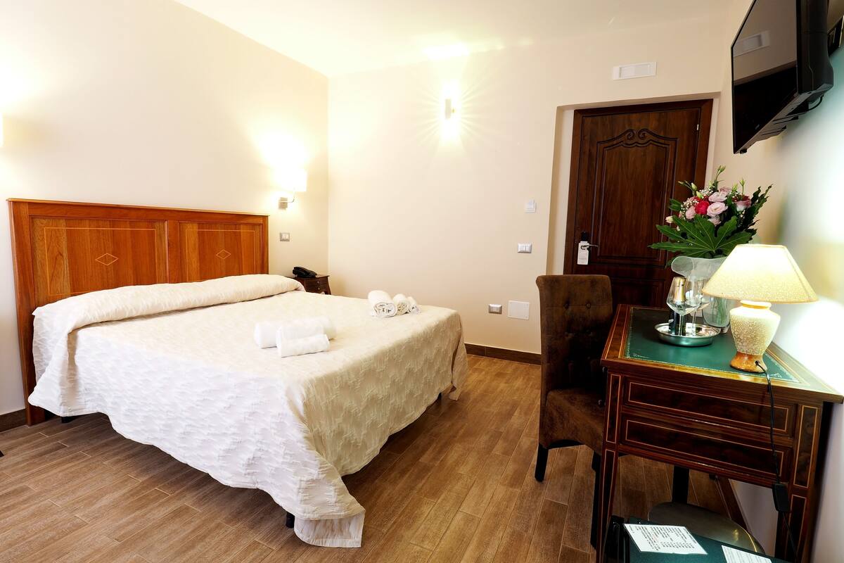 Habitación de hotel con cama de cabecera de madera, un escritorio con lámpara, una silla y un televisor de pantalla plana montado en la pared.