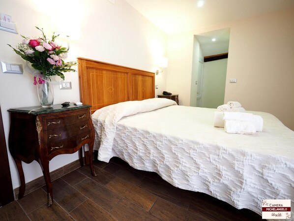 Une chambre d’hôtel avec une tête de lit en bois, une table de chevet ornée d’un vase de fleurs, un lit avec une literie blanche et une petite table de nuit avec un téléphone.