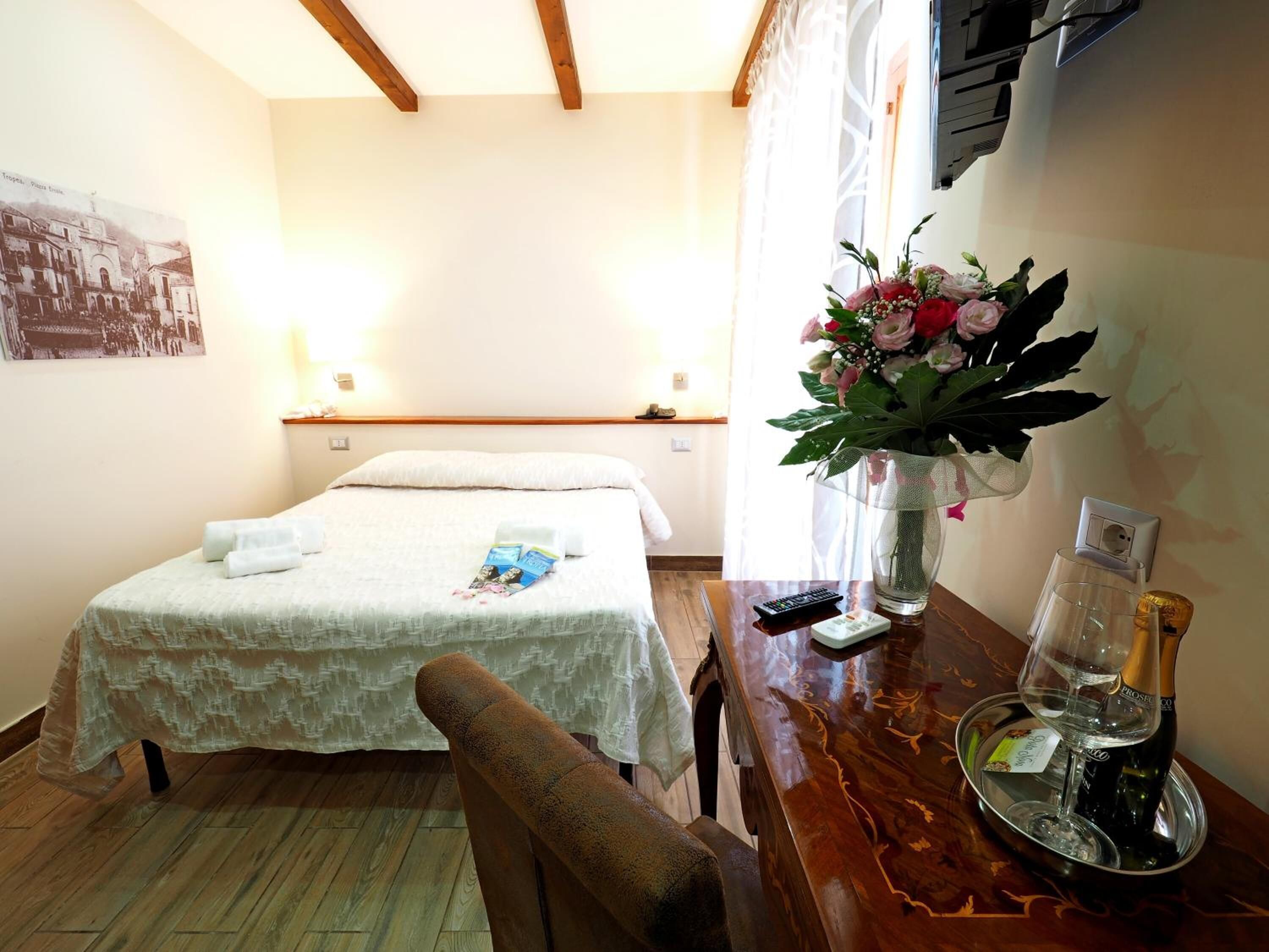 A hotel room with a bed, a wooden chair, a table with a vase of flowers, a bottle of champagne, and a remote control.