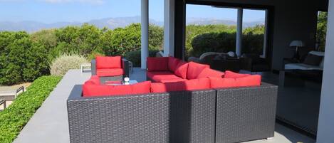 A modern outdoor seating area with a large sectional sofa and a dining table, surrounded by greenery and a view of distant mountains.