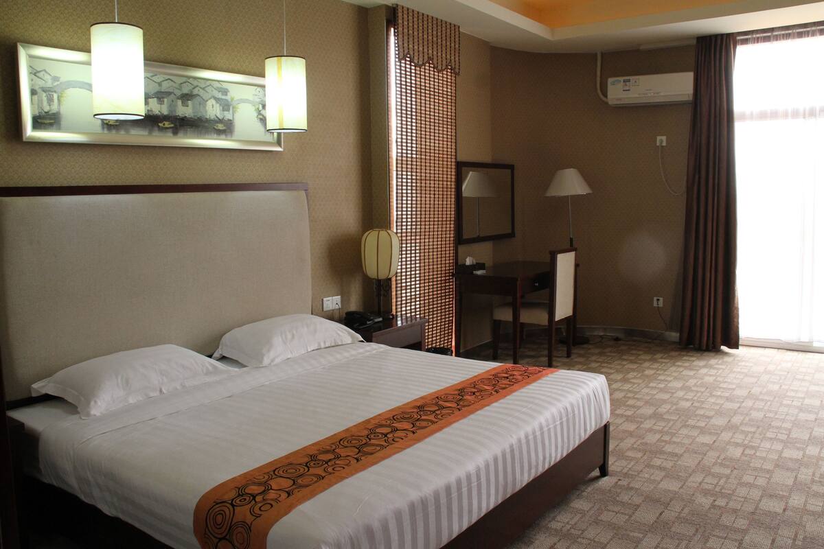 A hotel room with a large bed, a desk, and a chair. There is a window with curtains, an air conditioning unit, and a lamp on the desk.