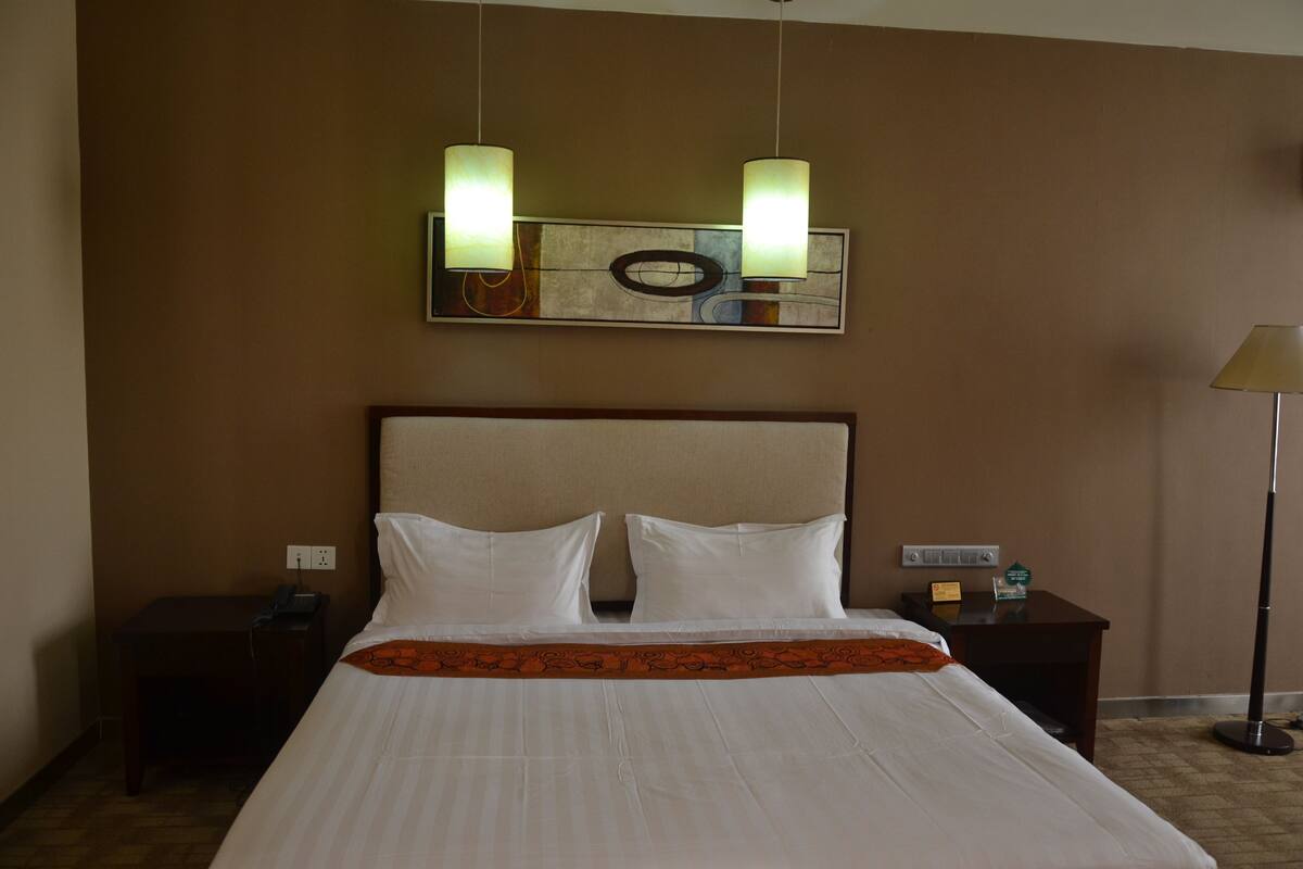 A hotel room with a large bed, two bedside tables, a nightstand, a lamp, and a wall-mounted artwork.