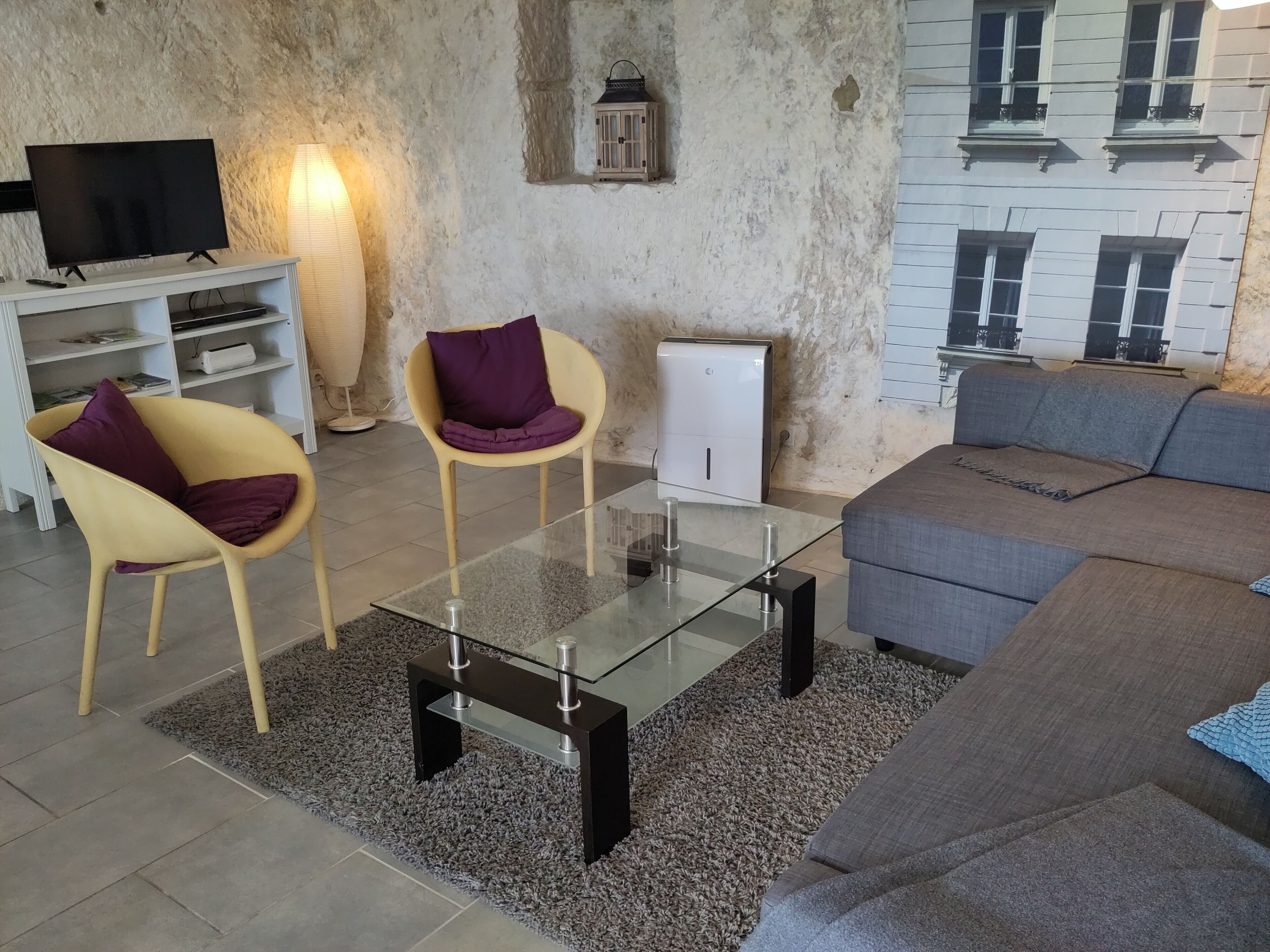 A modern living room with a glass coffee table, two yellow chairs with purple cushions, a grey sofa, a white cabinet, and a television on a white stand.