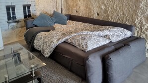 Une chambre avec un mur en pierre, un lit recouvert d’une couvre-lit à motifs et une table basse en verre.