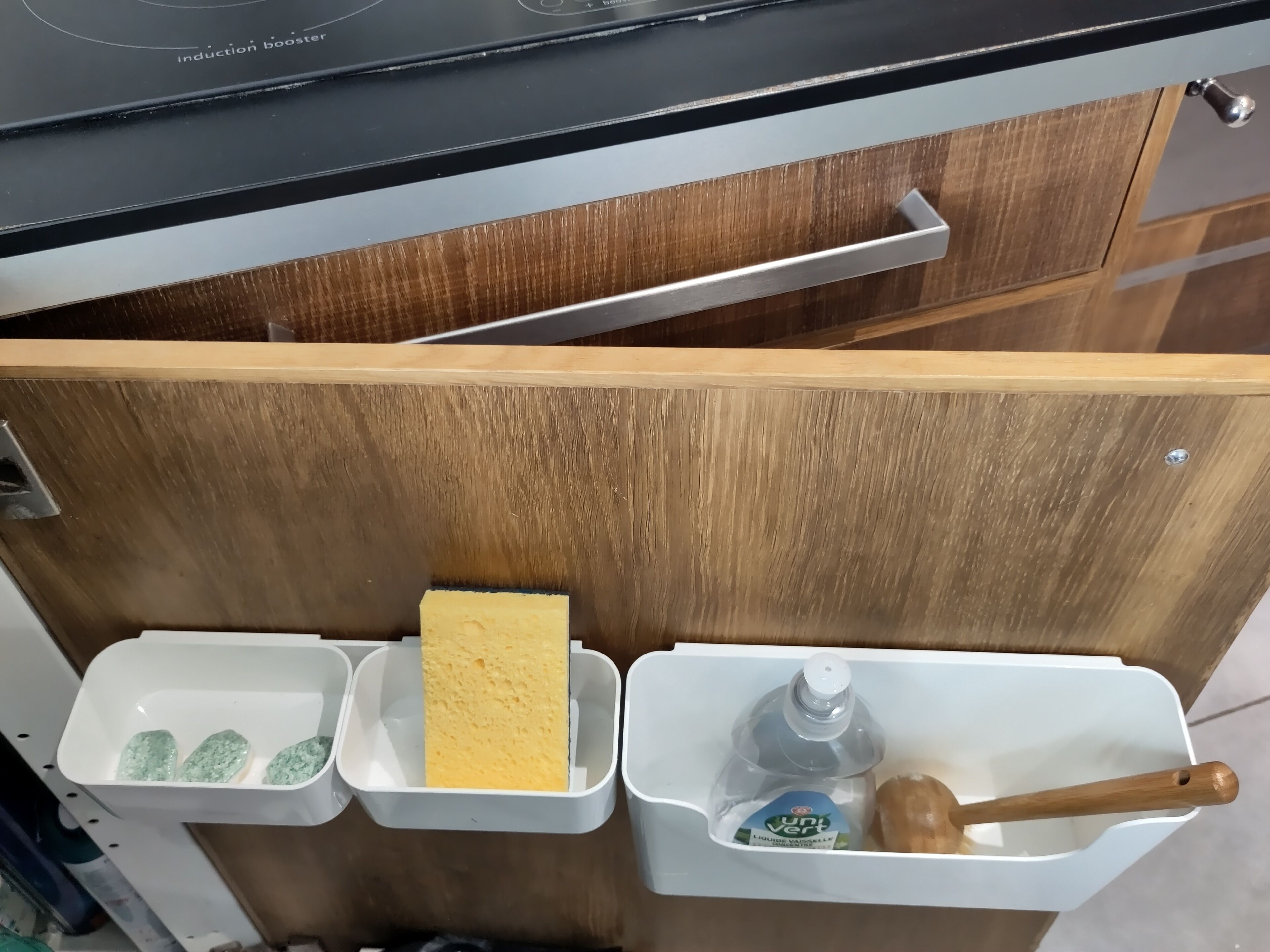 Un cajón de cocina con un tirador de madera, que contiene una esponja, una botella de agua y una cuchara de madera.