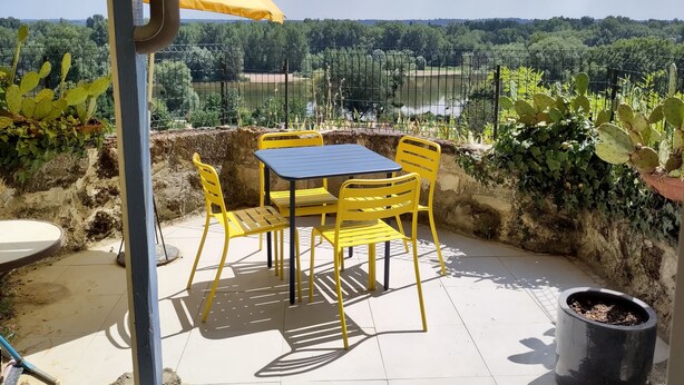 Une terrasse avec des chaises jaunes et une table, un parasol jaune, et une vue sur un lac et des arbres.