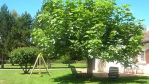 A backyard with a large tree, a swing set, and a patio area with outdoor furniture. - GÎTE LE CATALPA REGION OF THE HIGH LIMOUSIN (Saint-Georges-Les-Landes)