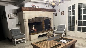 A cozy living room with a fireplace, two armchairs, a coffee table with a chessboard, and a door with glass panels. - GÎTE LE CATALPA REGION OF THE HIGH LIMOUSIN (Saint-Georges-Les-Landes)