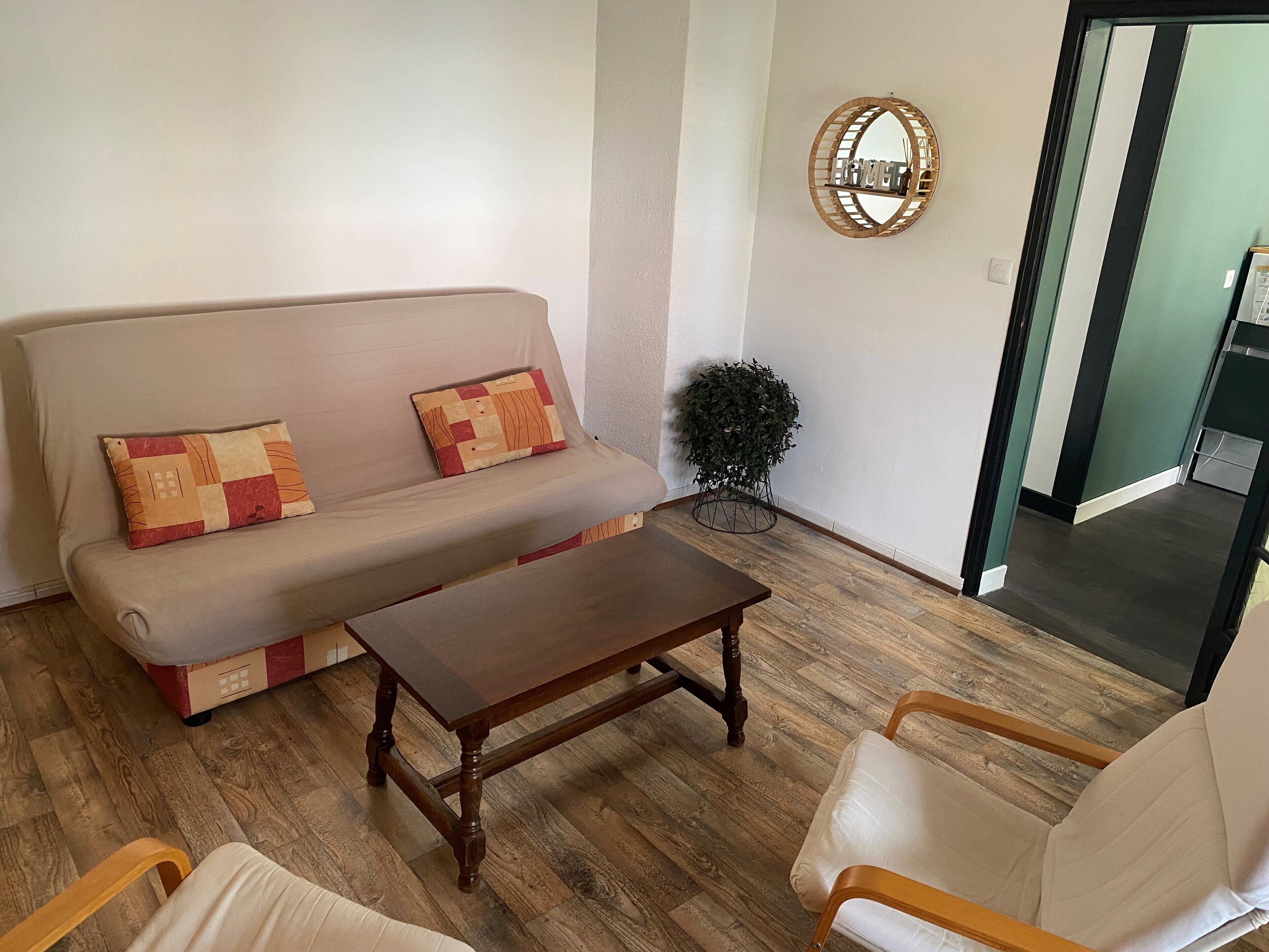 Ein Wohnzimmer mit einer Couch, zwei Sesseln, einem Couchtisch, einer kleinen Topfpflanze und einem Spiegel an der Wand.