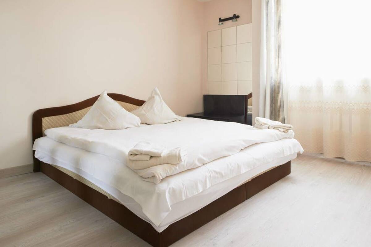 A neatly made bed with white linens and pillows, a wooden headboard, a small bedside table with a lamp, and a large mirror on the wall.