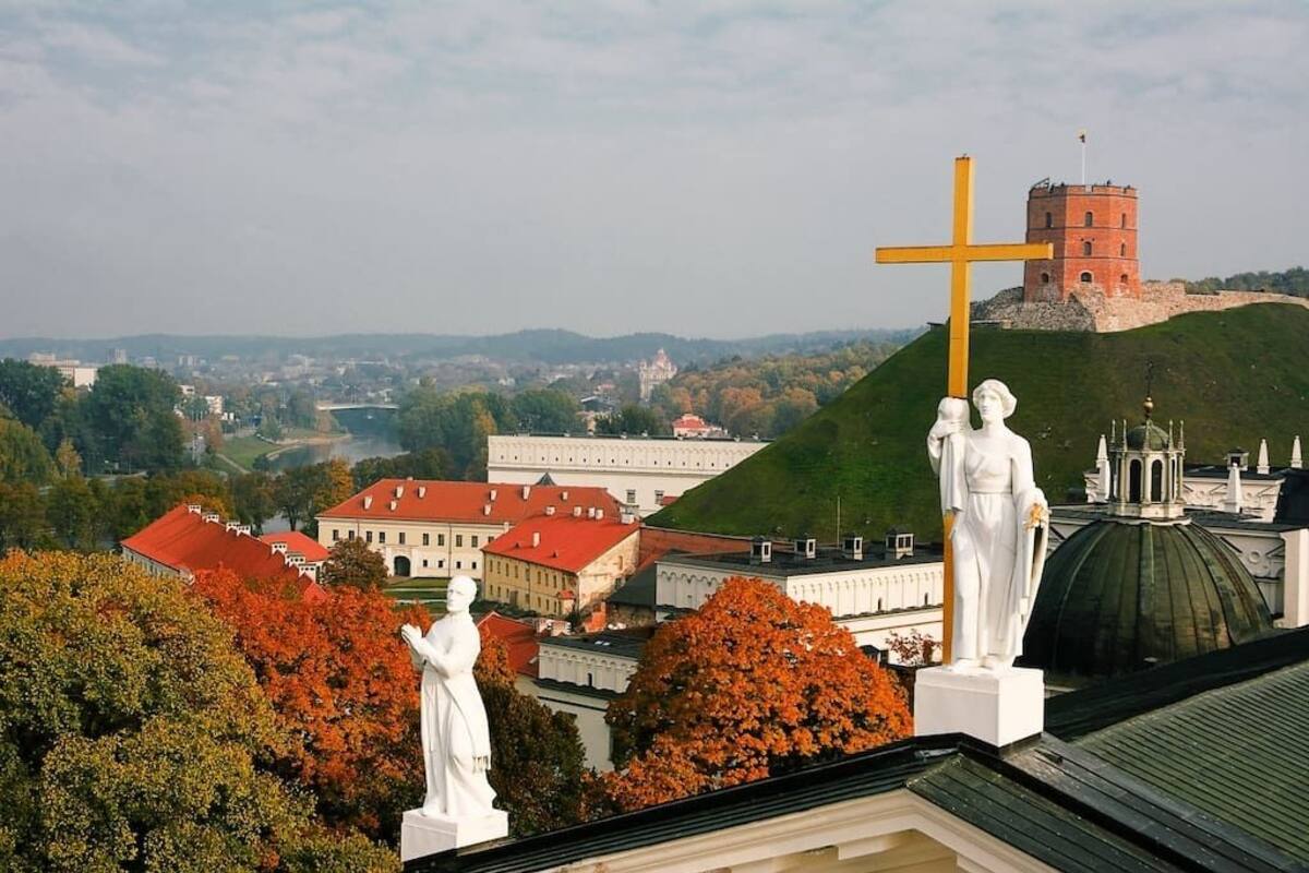 A view of a cityscape with statues, a prominent yellow cross, and a castle-like structure in the background.