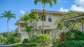A two-story house with a balcony, surrounded by lush greenery and palm trees.