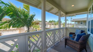 A covered porch with a wicker chair and cushions, a white railing, and a view of neighboring houses and palm trees.